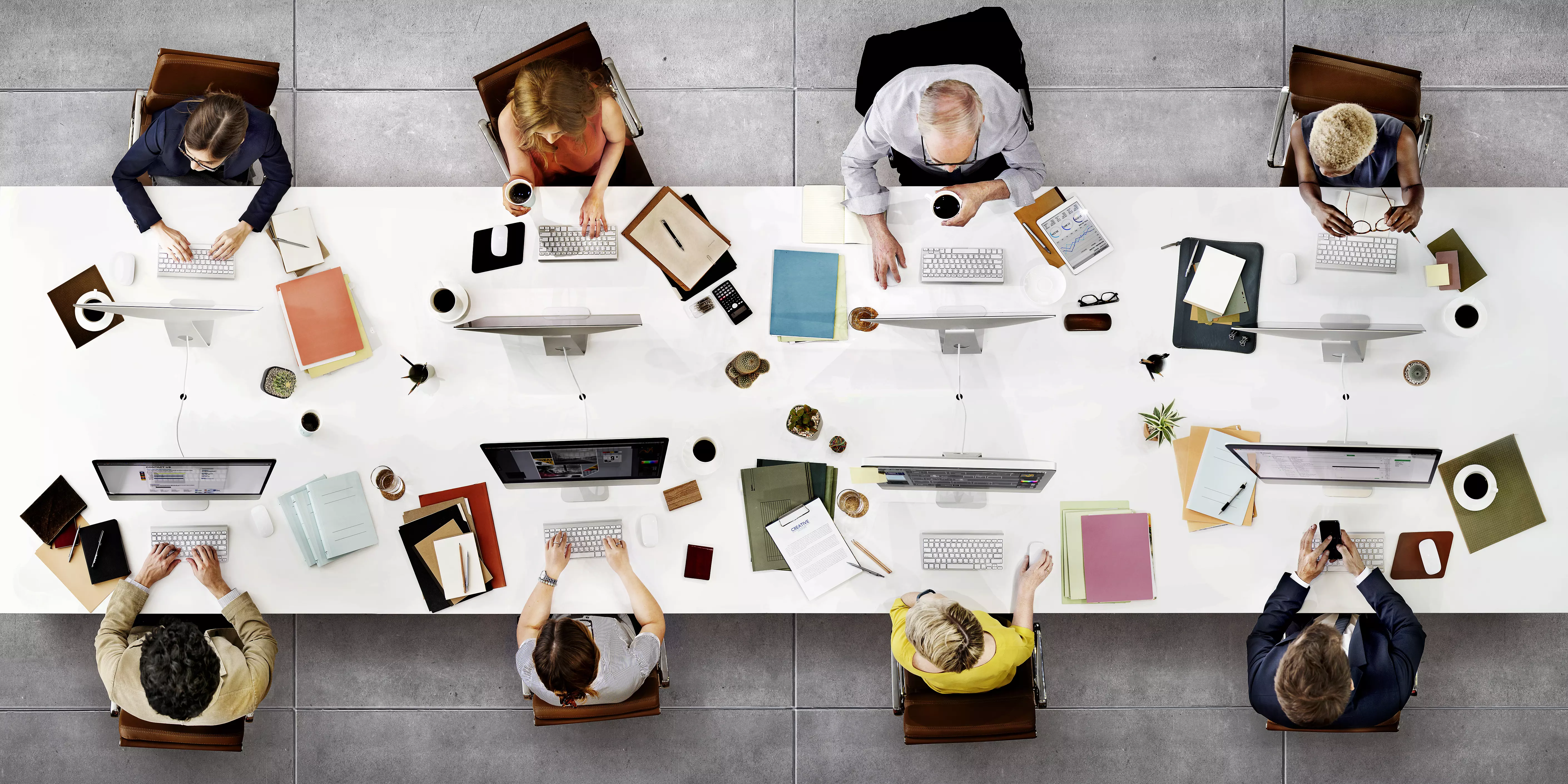 Fotografia przedstawiająca od góry stół wokół którego pracuje 8 osób. Na stole monitory, klawiatury, dokumenty, filiżanki.